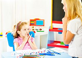 Woman sitting in front a girl places her fingers near her mouth to show speech during a lesson.