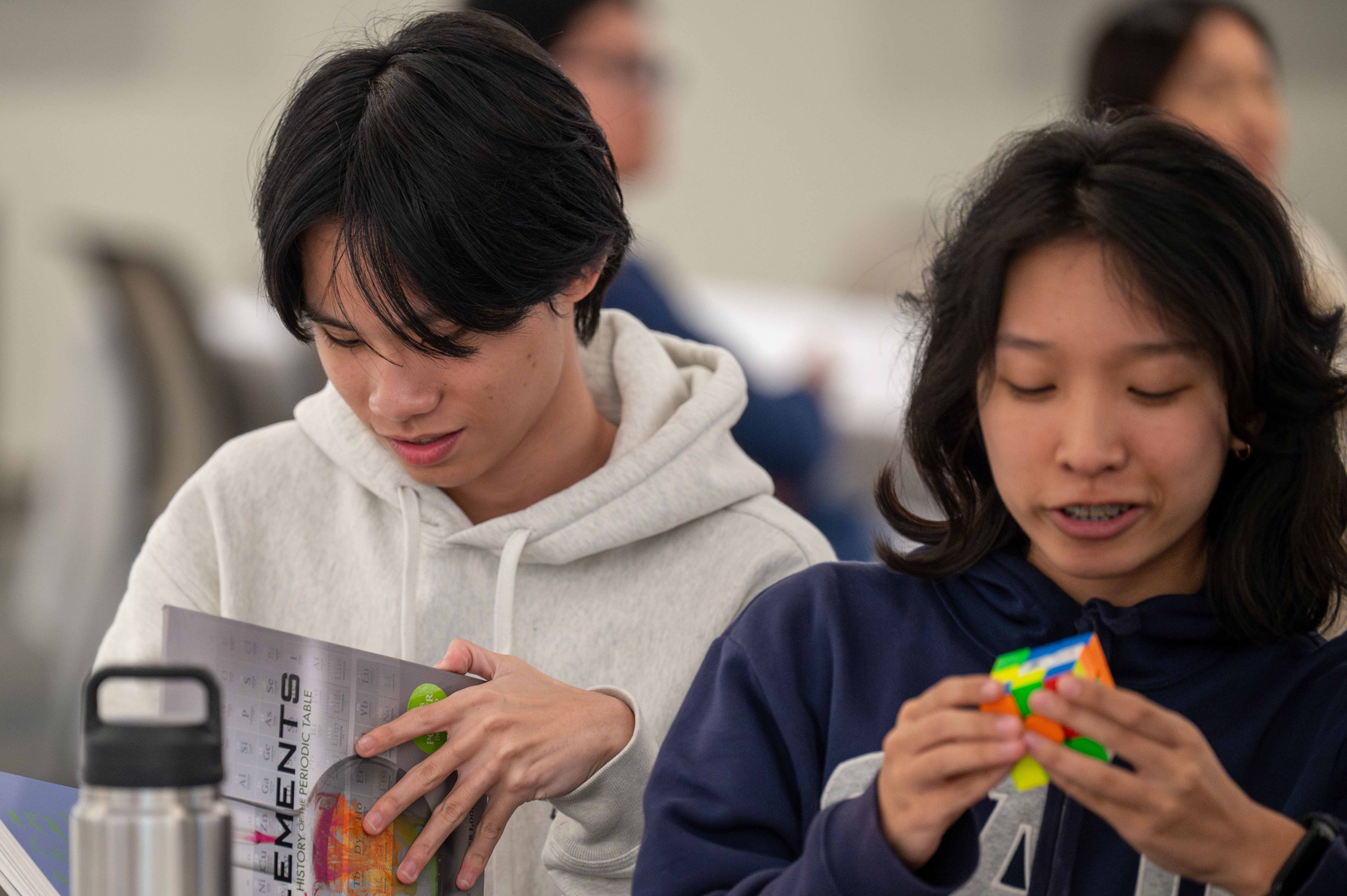 One student reads through a textbook he just won and another attempts to solve a custom Math Meet Rubik's Cube